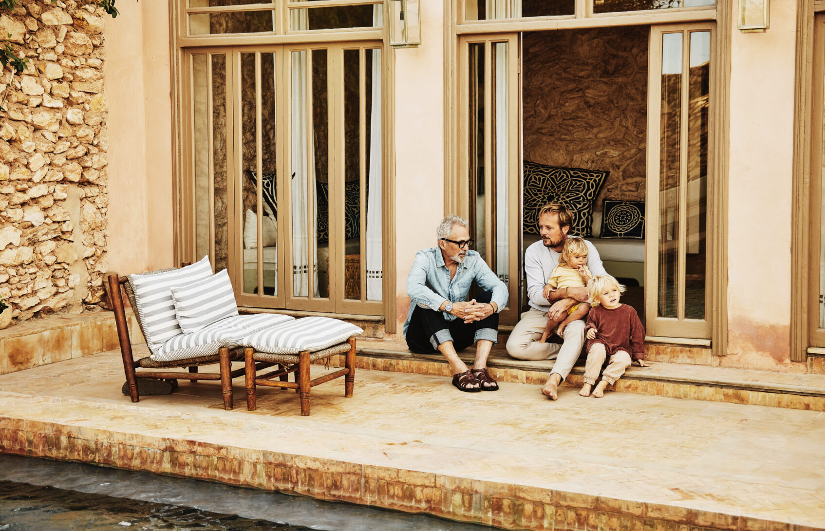 Father and adult son in discussion while relaxing with family on poolside terrace of villa
