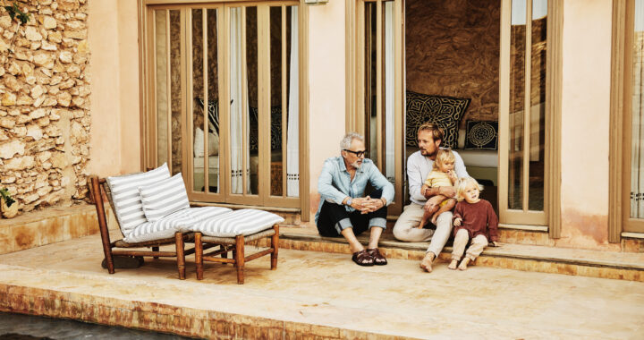 Father and adult son in discussion while relaxing with family on poolside terrace of villa